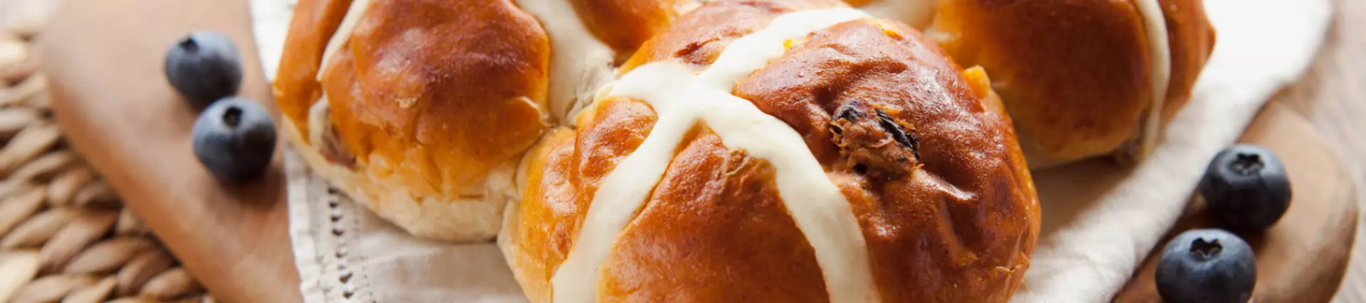 A wooden tray full of hot cross buns, blueberries are scattered around the edge