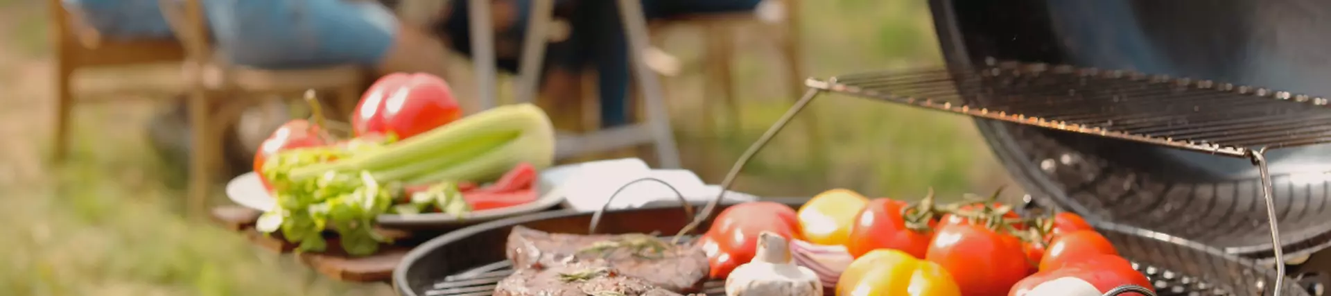 A BBQ with steak and veggies cooking in the foreground. In the background a bench with friends